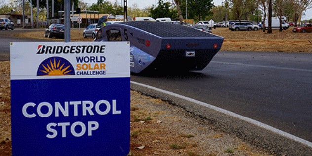 A black, sleek, aerodynamic, futuristic vehicle drives along a road past a sign saying Control Stop.