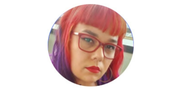 A photo of Susan Wood looking at the camera with red glasses and pink fringe fading to longer purple hair.