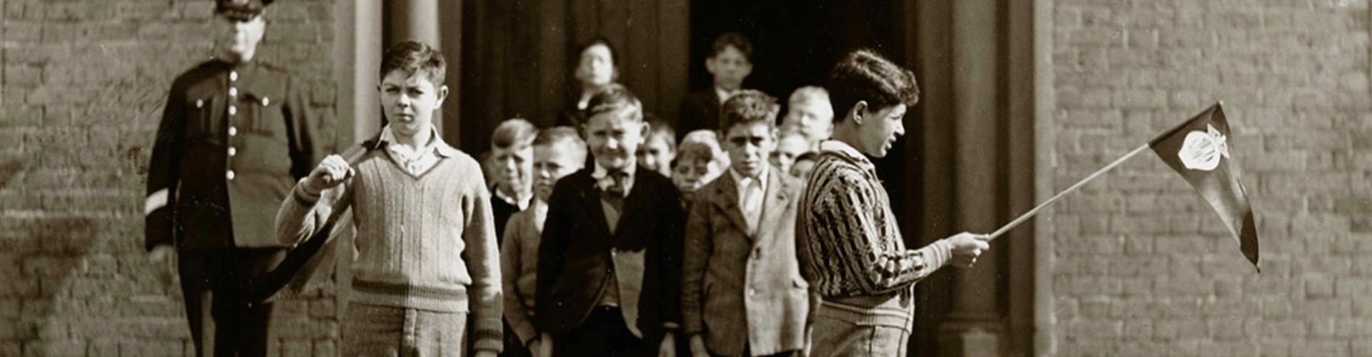 NRMA 1930s School Safety Patrols - Flags black and white photo showing two boys holding flags while a larger group of boys waits for the signal that it is safe to cross the road
