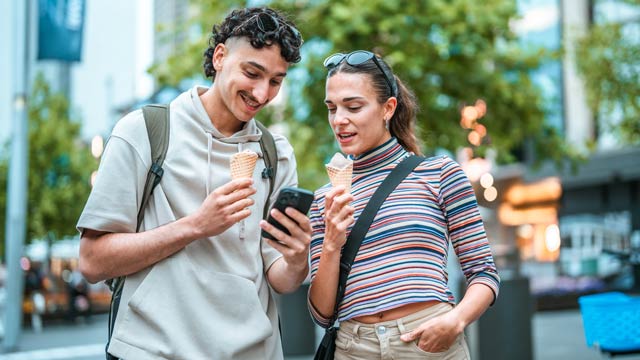 A couple eating icecream is a leaefy street looking at a phone