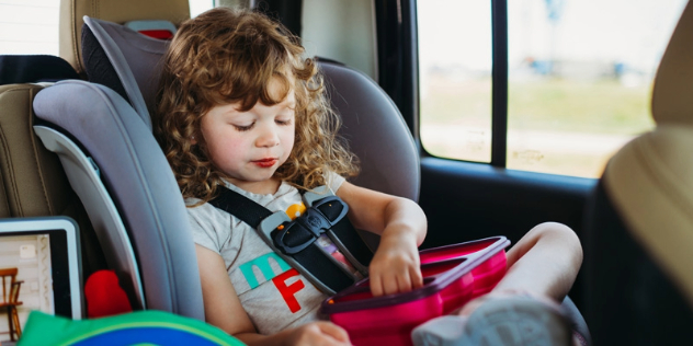 small girl in a child seat eats snacks from a red lunchbox