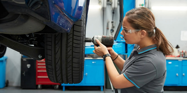 Side view of a lady holding an impact drill and removing the wheelnut from a tyre while wearing safety glasses.