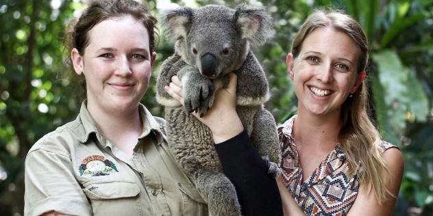 female sanctuary staff member holds koala between herself and a female visitor with long blond hair for a photo with a koala