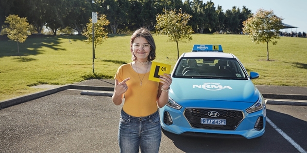 young female learner driver in an orange shirt and jeans stands next to an NRMA learner car and points to an L plate in her hand