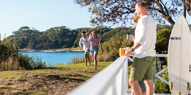 parents watch two children play on the grass in front of their holiday cabin next to the beach