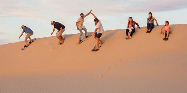 people preparing to surf down sand dunes, four are standing and three are sitting