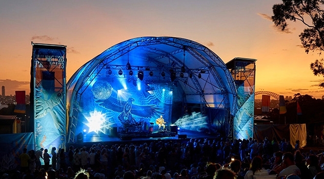 Twilight view of a band playing on the stage lit in blue light with a sunset and Sydney harbour bridge in the background