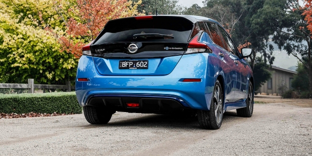 Rear view of a compact blue car parked in a driveway, with trees on the left hand side.