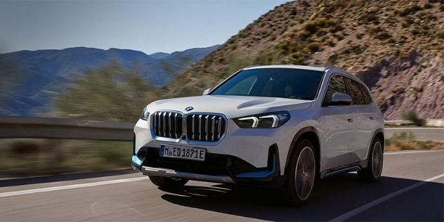 A front-angle view of a white BMW iX1 driving along a windy road adjacent to a rocky mountain.