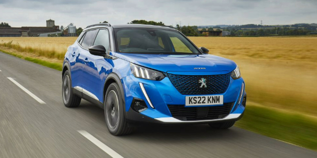 A blue Peugeot E 2008 driving along a road adjacent to dry grassland. 