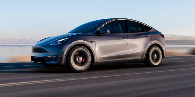 Side view of a gold Tesla Model Y driving along a dirt road adjacent to a body of water.