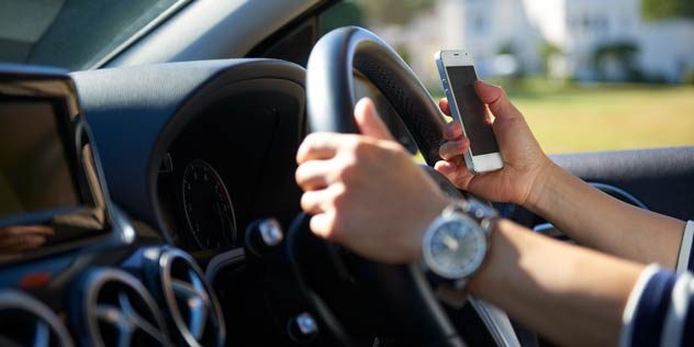 Two hands in a car, one holding a mobile phone and the other on the steering sheel
