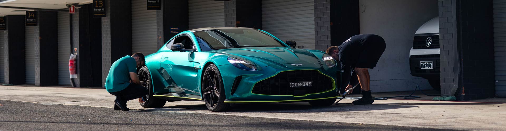 Two people check the tyres on a turquoise Aston Martin in front of parking area
