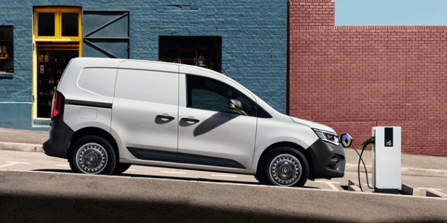 A white Renault Kangoo E-Tech parked on an inclined street and plugged into a charging station