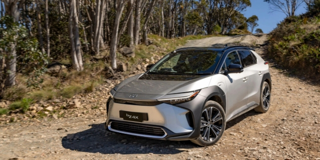 Front view of a silver Toyota bZ4X parked at an angle on a dirt road, with tall trees beside it.