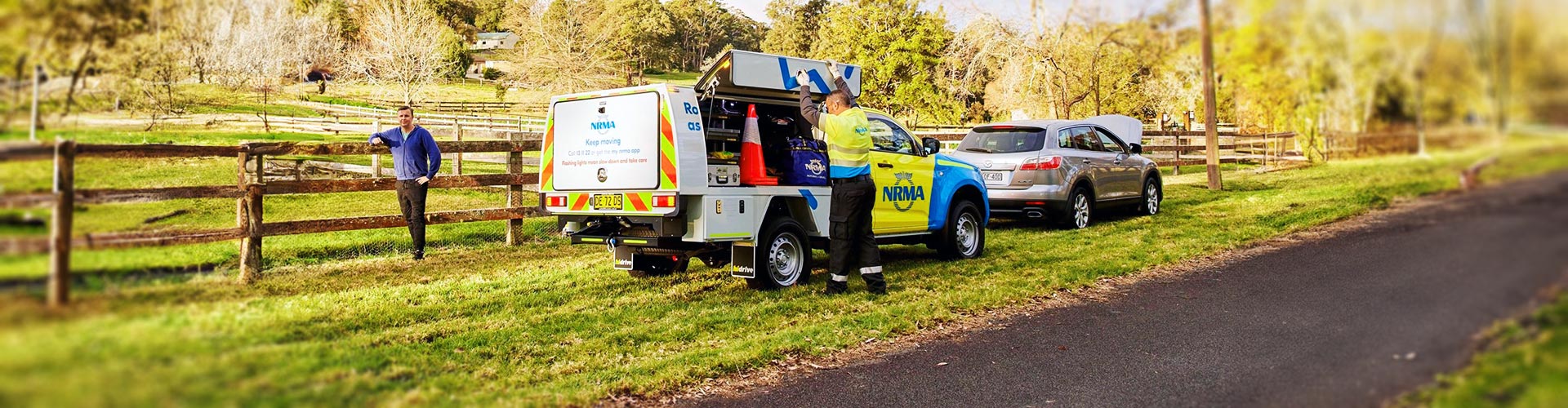 A man leans on a fence while roadside assistance attends to his car