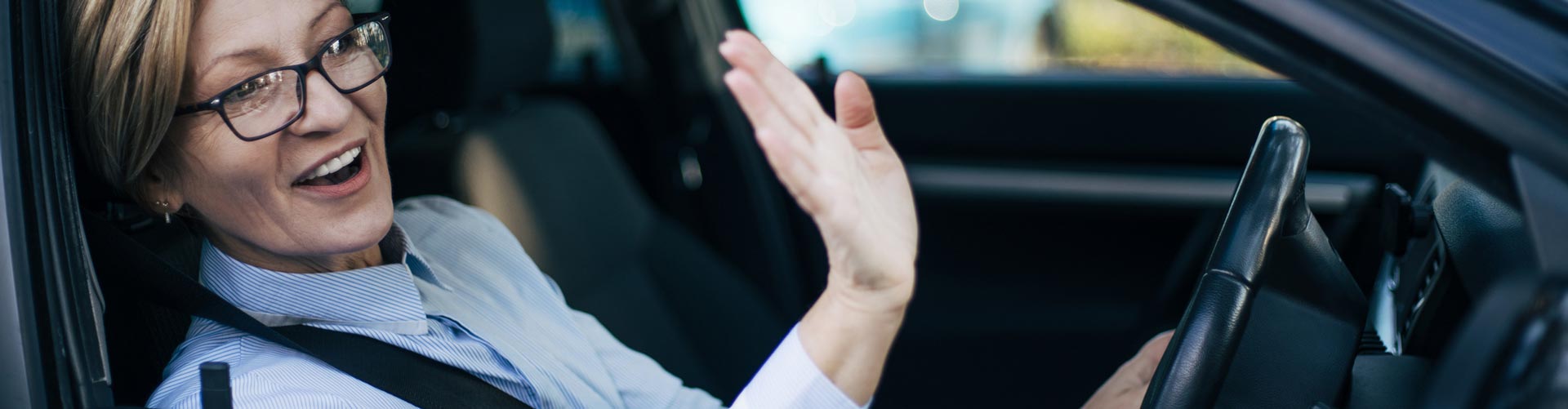 Middle-aged woman sitting in driving seat of car and waving to someone outside the car