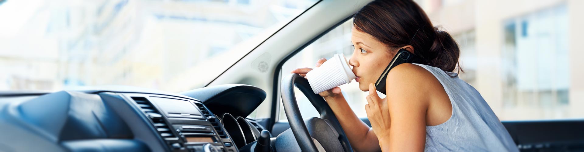 A young woman talks on the phone and holds a coffee cup while at the wheel of a car