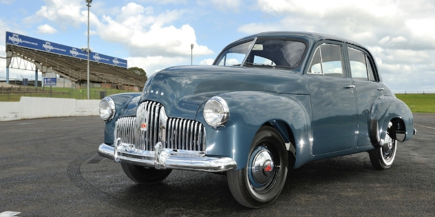 Front-angle view of a blue vintage car parked on a black tar surface.