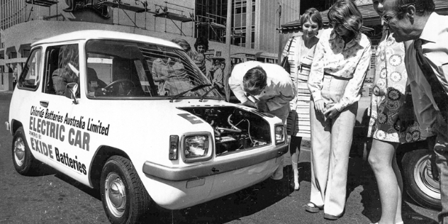 A black and white photo of a group of people standing around the front of a vintage car.