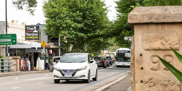 Front view of a white Nissan traveling along a city street with shops on one side and stone pillar on the right-hand-side foreground.