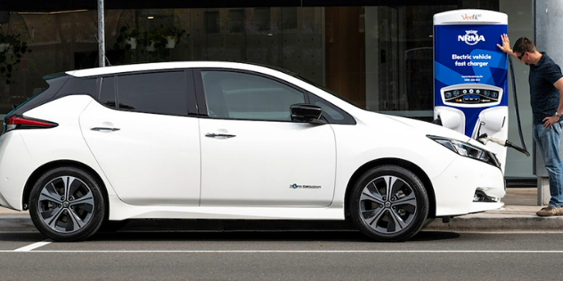 A man leaning on an NRMA fast charger, while standing in front of a his white car as it charges.