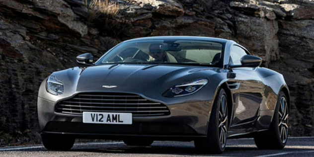 Front view of a grey, two-door Aston Martin travelling along a road with a rock face in the background.