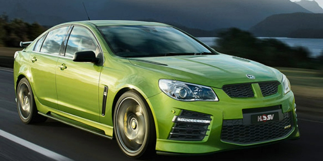 Front view of a sporty green sedan travelling along a road, with mountains and a river in the background.