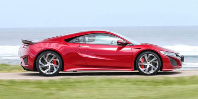 Side view of a sleek and low, red, two-door car travelling adjacent to the beach.