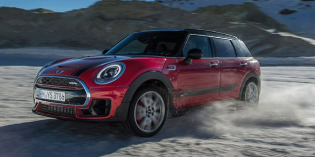 A sporty red Mini Cooper taking a sharp turn and kicking up dirt on sandy outdoor terrain.