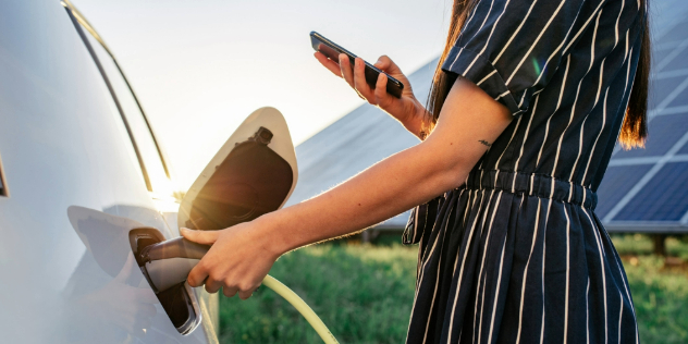 A woman looking at her phone while holding a charger plugged the back of her car, with slanted solar panels behind her.