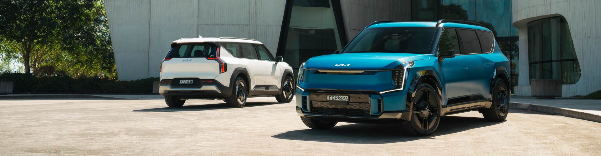 Two blue and white 2023 Kia EV9s parked in front of an angular building