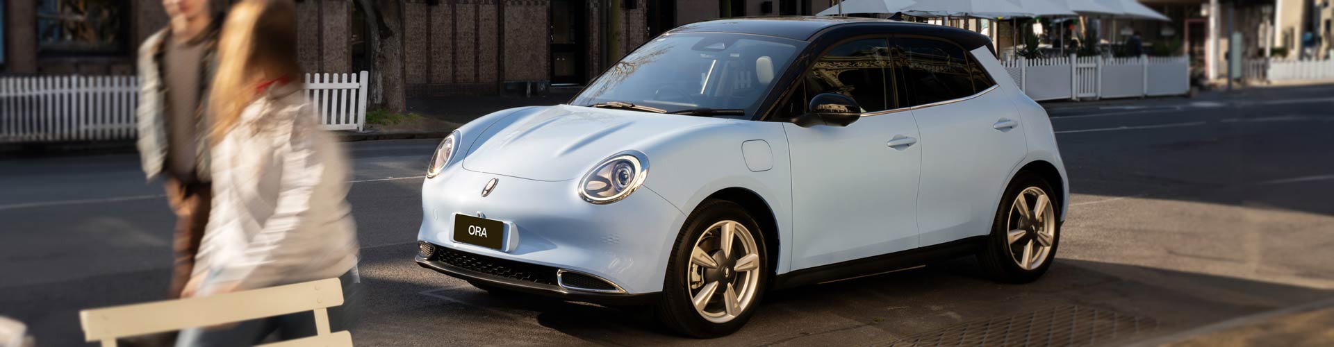 A blue GWM Ora electric hatch drive down a city street next to a walking couple