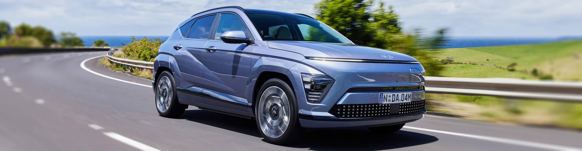 A blue 2024 Hyundai Kona EV driving on a country road with the ocean behind