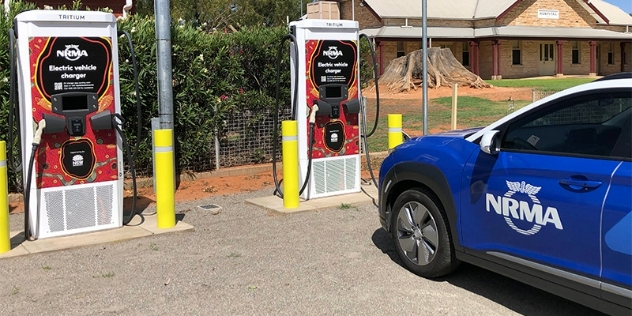 An electric vehicle branded in the blue and white NRMA colours, parked in front of a charging station which has been designed with an intricate and colourful piece of indigenous art.