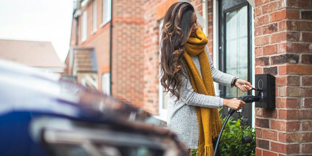 A woman with long hair wearing an orange scarf charges her EV at home