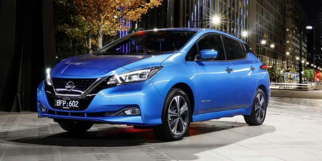 A blue Nissan Leaf, four door hatchback parked in the pavement at night in along a city street with a row of street lights in the background.