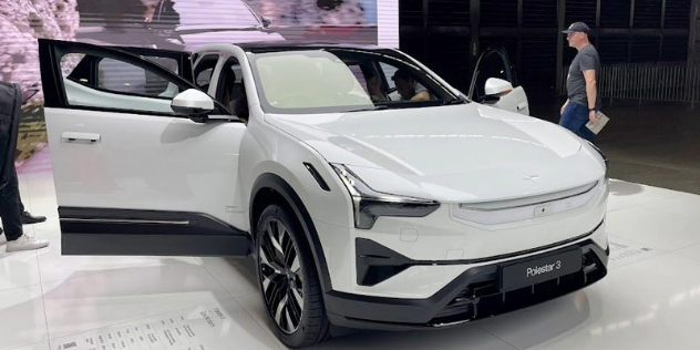 Side-angle view of a white Polestar 3 electric vehicle parked in a showroom with its doors open and people walking around and viewing the car.