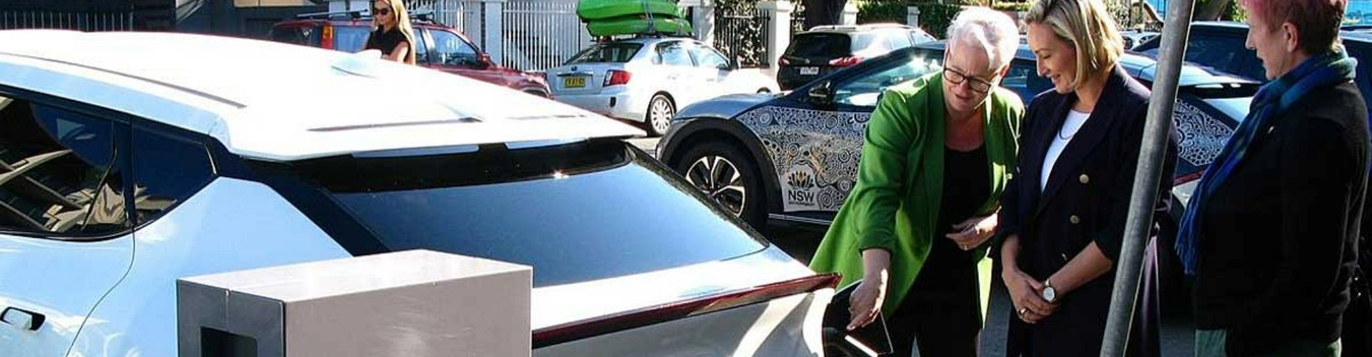 Three women standing around a car that's being charged by a kerbside charger.