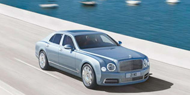 A silver sedan travelling along a road next to a harbour.