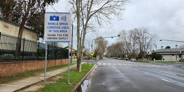 Front view of a mobile speed camera sign board on the pavement, to the left of a road on a dull day.