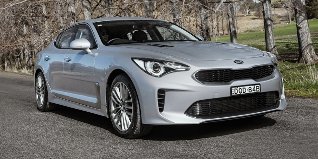 Front view of a silver Kia Stinger travelling along a road with tall trees in the background.