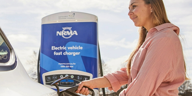 A woman plugging a charger into the back of her car under a sign that says, NRMA electric vehicle fast charger.