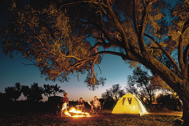 Father and children sit by a fire next to a tent under a gum tree at night