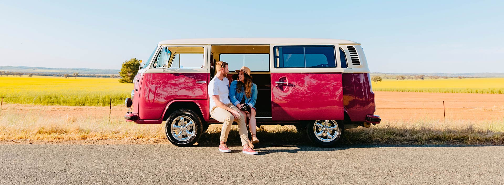 A couple sits in a red and white Kombi in front of a canola field