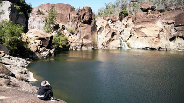 A woman sits facing the orange rock fall acorss Macintyre Falls waterhole