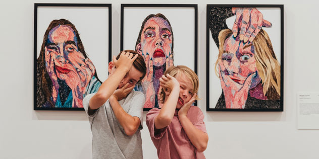 Two children squish their faces in front of some paintings
