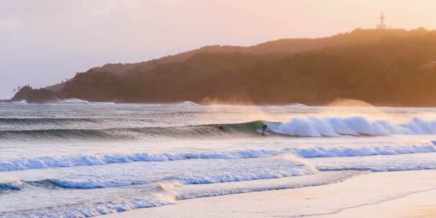 A surfer on a wave in front of Byron Bay lighthouse at sunset