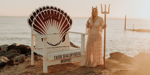 A woman in a flowy white dress stands next to a Scallop Fiesta sign