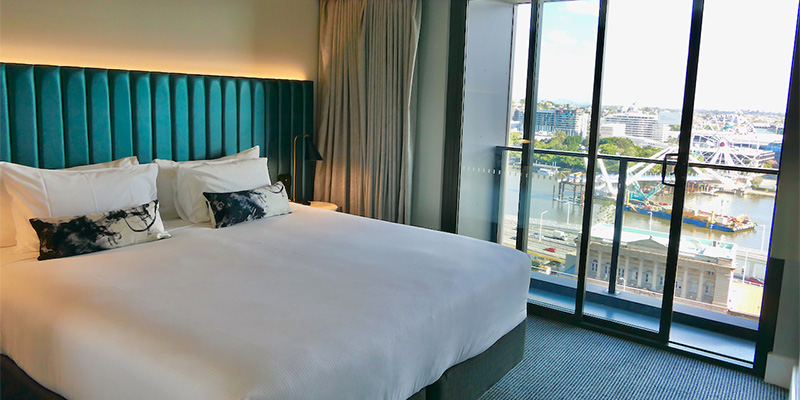  A king size hotel room bed, with a blue upholstered headboard, accent pillows, and a balcony overlooking a city riverfront.